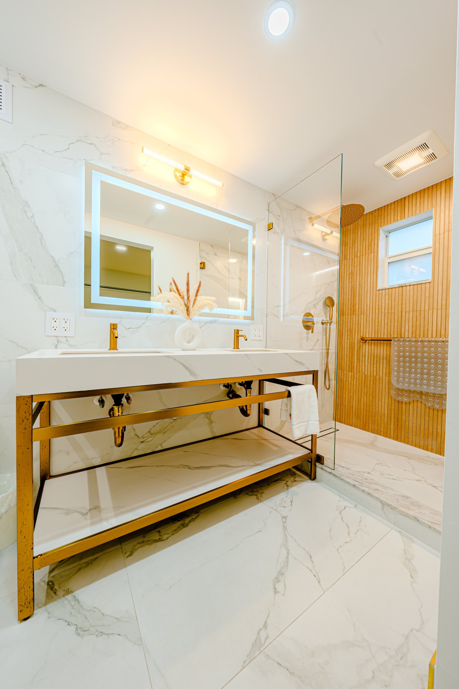 A modern bathroom with a marble-tiled floor and walls, gold fixtures, and a glass-enclosed shower.