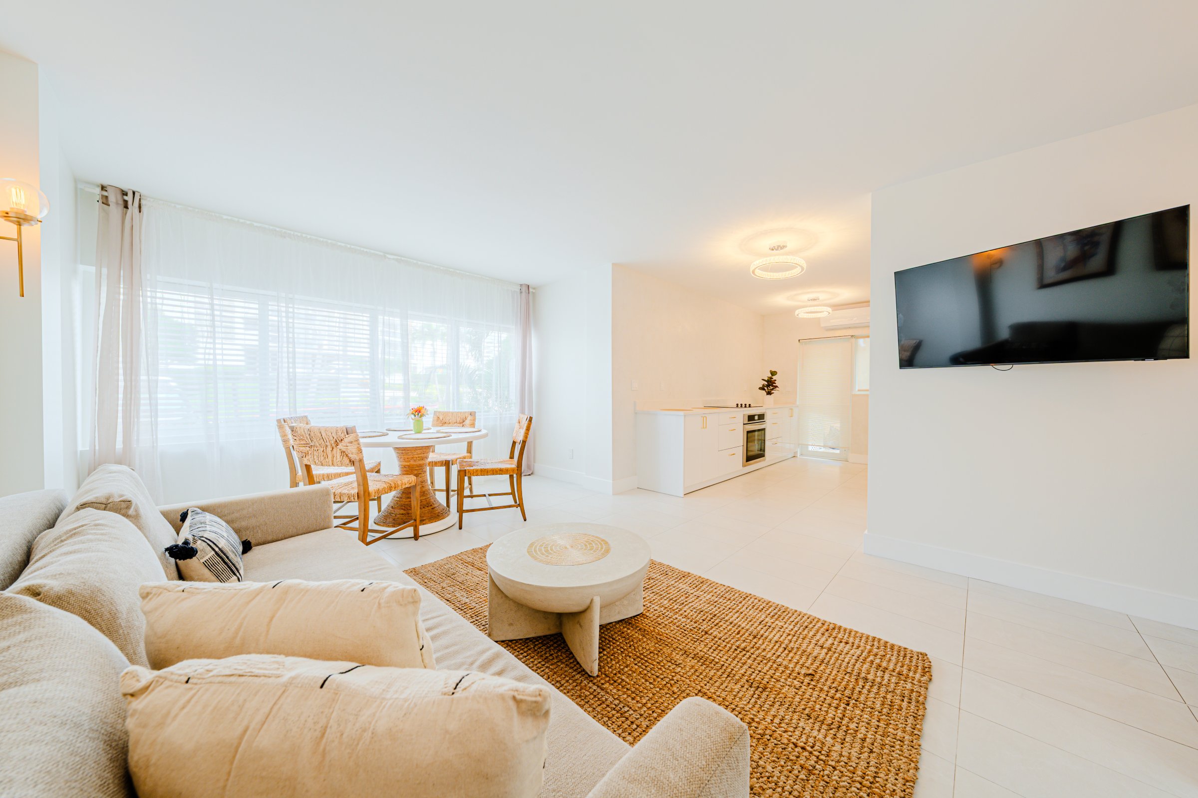 A bright and airy living room with a sofa, TV, dining table, and kitchenette.